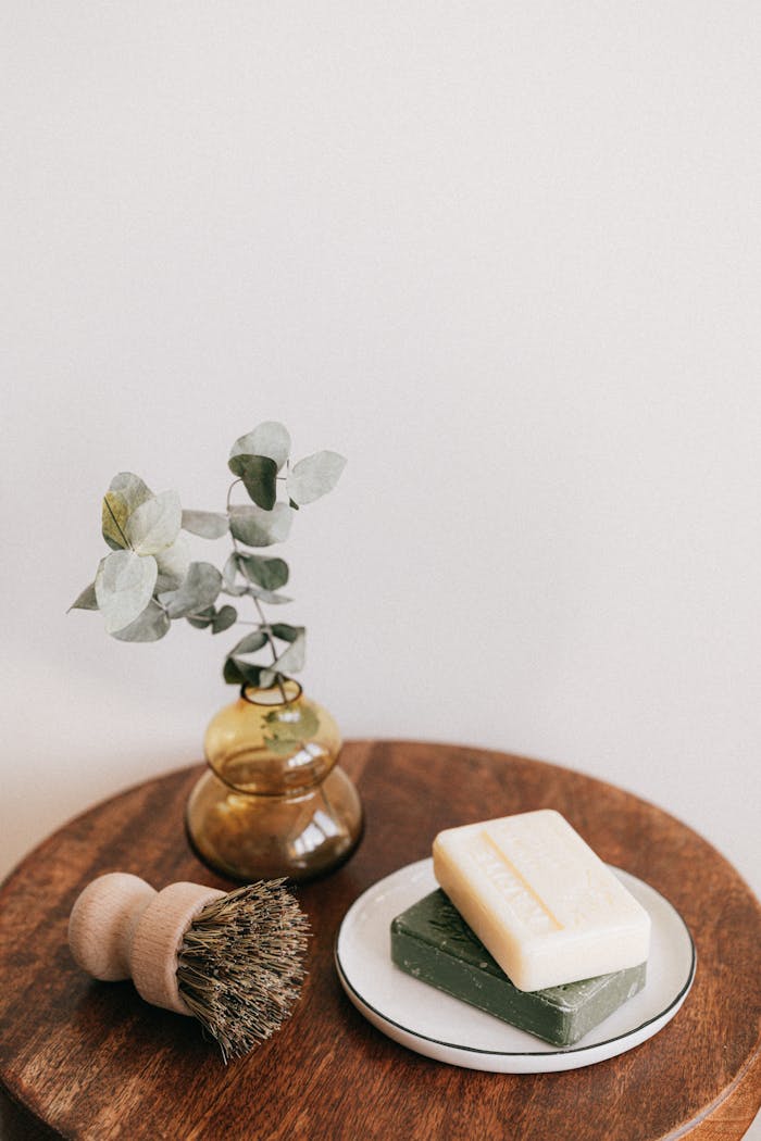 From above composition with assorted organic soaps on white ceramic holder and shaving brush with wooden handle near yellow transparent vase with plant on wooden round table against white wall